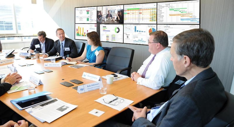5 موظفين يجرون مناقشة أثناء جلوسهم على طاولة في غرفة اجتماعات مع حائط فيديو يعرض تصورات البيانات