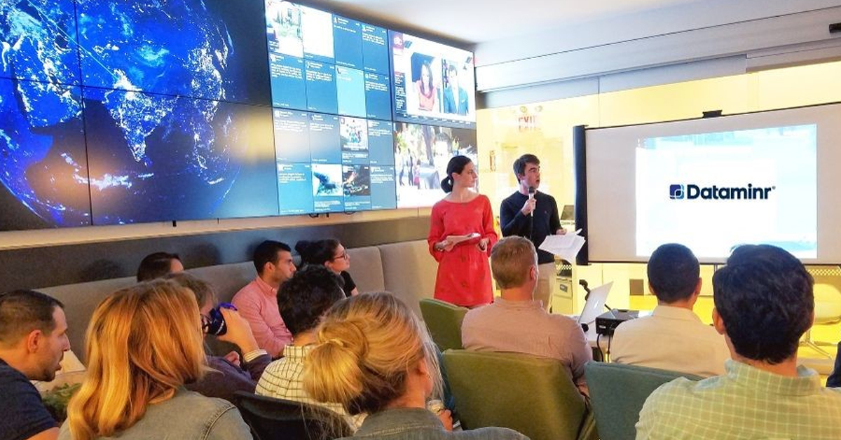موظفو Dataminr في غرفة اجتماعات يجلسون مقابل جهاز عرض ، وبجانب حائط فيديو مع تحديثات وسائل التواصل الاجتماعي والأخبار