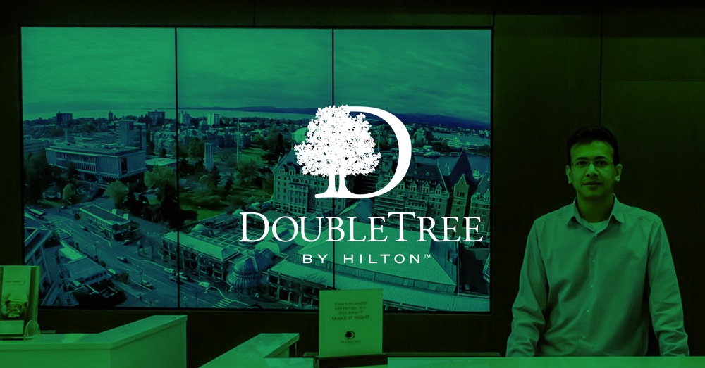 موظف استقبال DoubleTree by Hilton و 3 لوحات فيديو خلفه تعرض معالم من فيكتوريا ، كندا مع تراكب أخضر وشعار