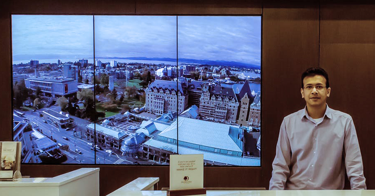 موظف استقبال دبل تري باي هيلتون و 3 لوحات فيديو خلفه تعرض معالم من فيكتوريا ، كندا