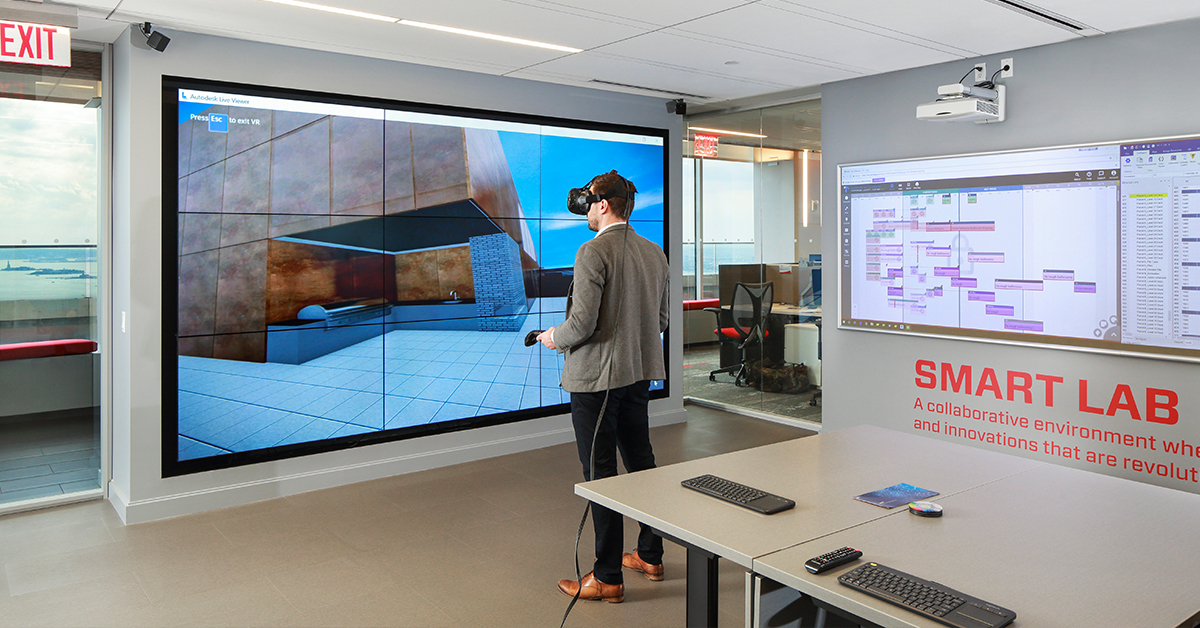 Suffolk's Smart Lab room with a desk and a man exploring virtual reality, and a video wall displaying what he sees virtually