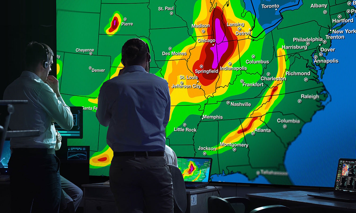 2 رجال في غرفة التحكم في مراقبة الطقس ، ينظرون إلى جدار فيديو كبير يعرض خريطة الطقس للولايات المتحدة الأمريكية
