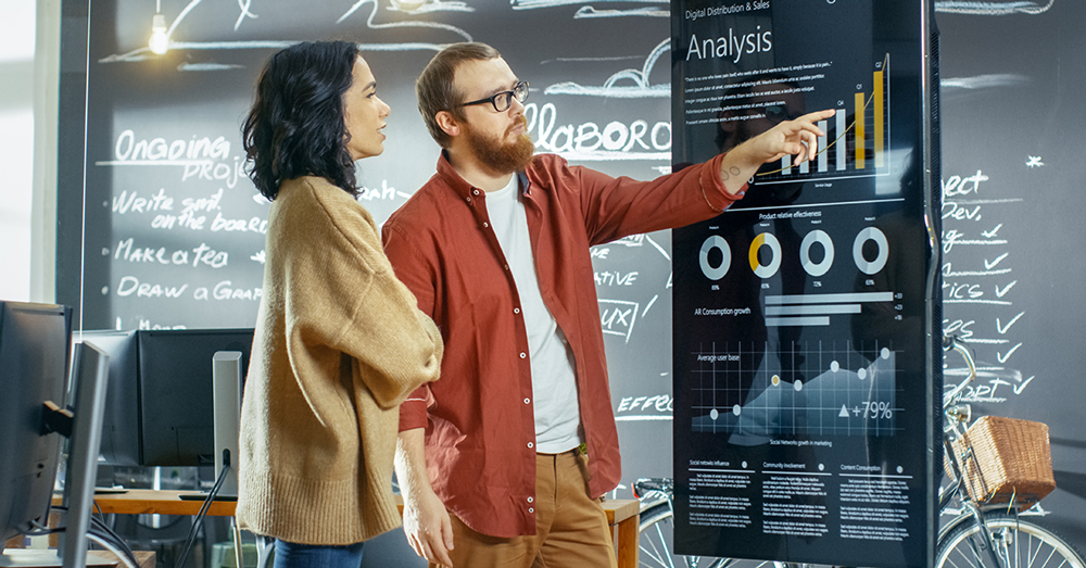 A man and woman discuss a data dashboard on a TV screen, with a chalkboard with project details behind them