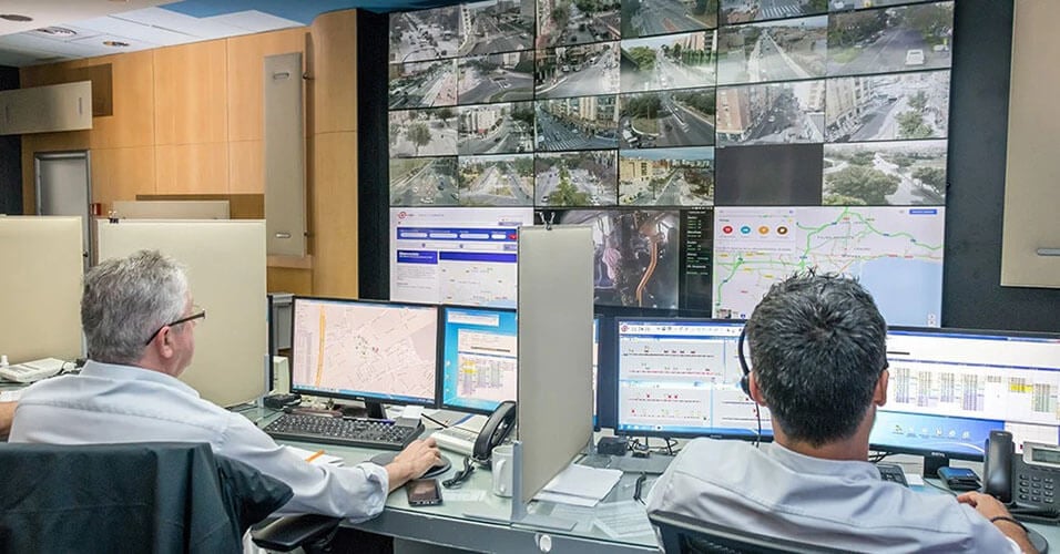 Two EMT employees monitoring transit operations through their workstations and a video wall displaying live camera footage, transit maps, and websites